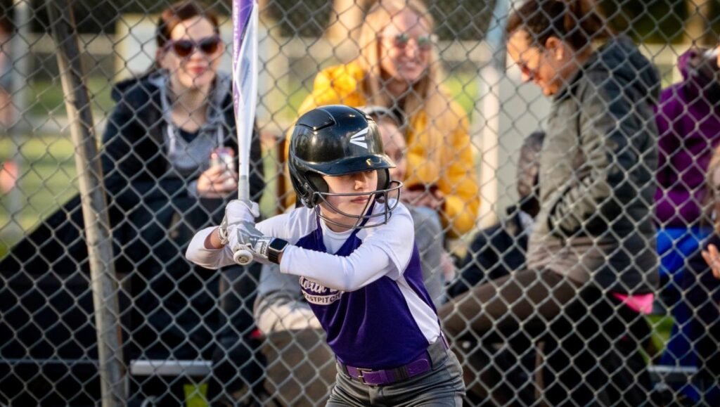 A young softball player wearing a black helmet and purple batter's jersey taking a powerful swing at the plate. The perspective is from behind a chain-link backstop net as spectators watch the game.