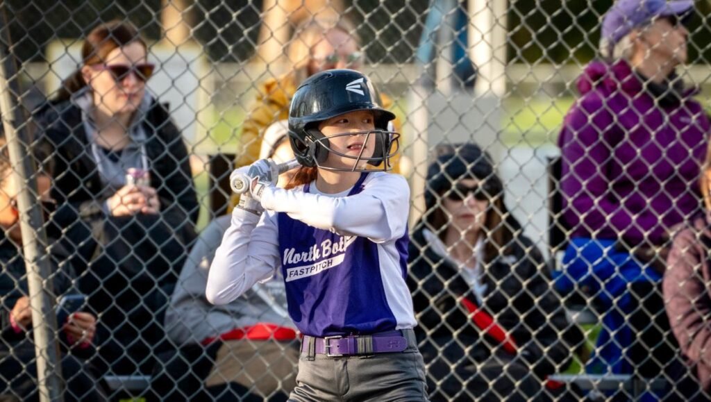 A baseball player prepares to bat while spectators watch from behind a chain-link fence.