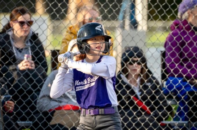 A baseball player prepares to bat while spectators watch from behind a chain-link fence.