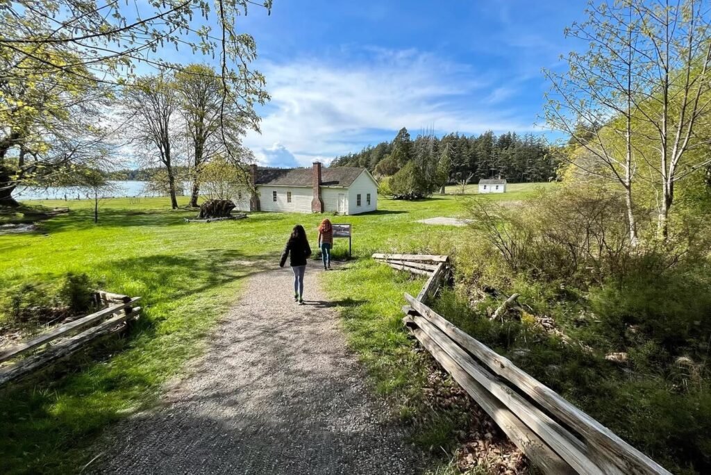 A gravel path lined with split-rail fences leads to a white church near the shore, with individuals strolling along the way.