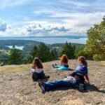 Bounce Around Individuals relaxing on a rocky outcrop overlooking a scenic lake and forest landscape under a sunny sky.