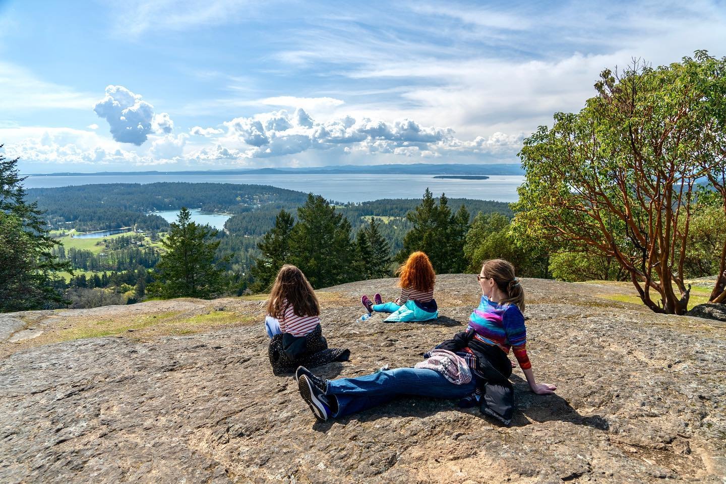 Young Hill at English Camp Individuals relaxing on a rocky outcrop overlooking a scenic lake and forest landscape under a sunny sky.