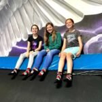 Three people sit on a trampoline in front of a large decorative angel wing. The image features a cosmic background painting at a trampoline park.