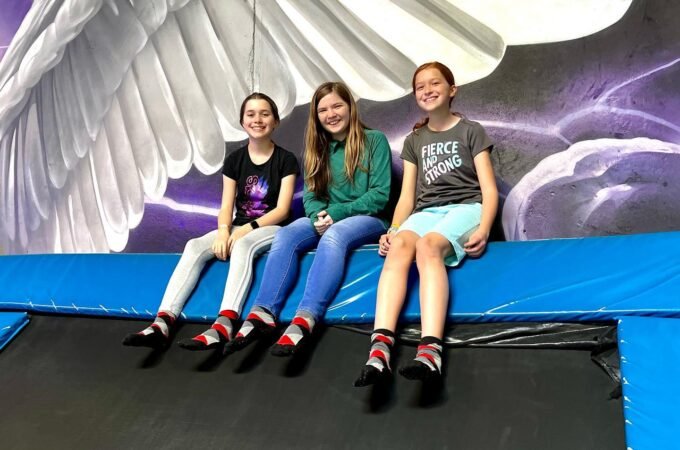 Three people sit on a trampoline in front of a large decorative angel wing. The image features a cosmic background painting at a trampoline park.