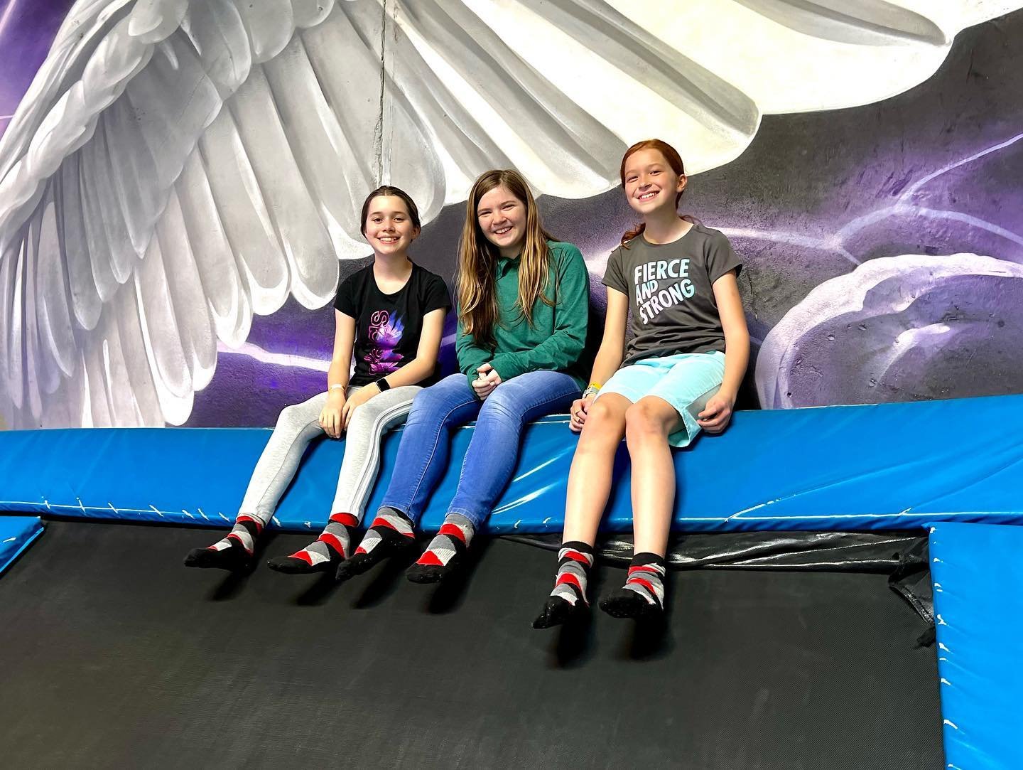 Bounce Around Three people sit on a trampoline in front of a large decorative angel wing. The image features a cosmic background painting at a trampoline park.