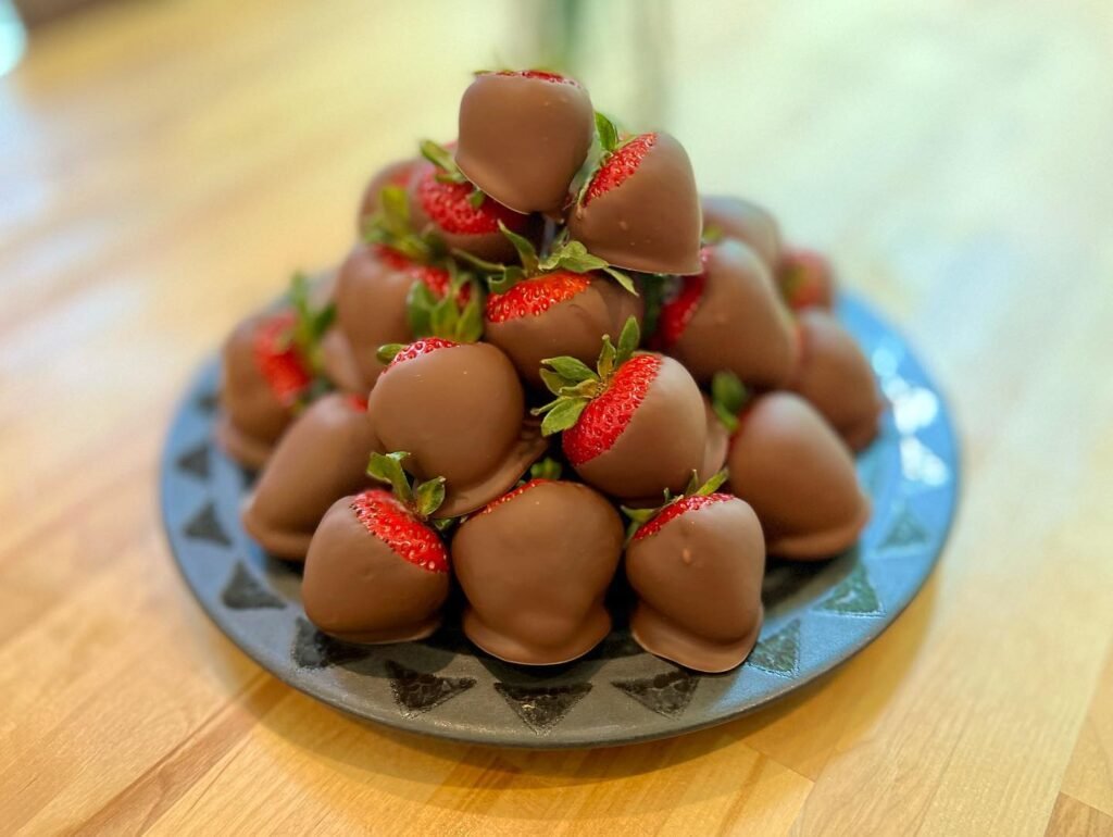 A pile of chocolate-dipped strawberries sits on a decorative blue plate on a wooden table in warm indoor light.