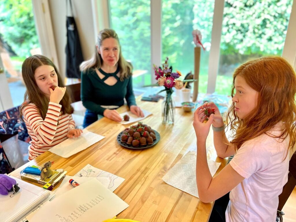 Three people are gathered around a wooden table, writing on paper. The table is adorned with a plate of chocolate truffles and a vase of flowers.