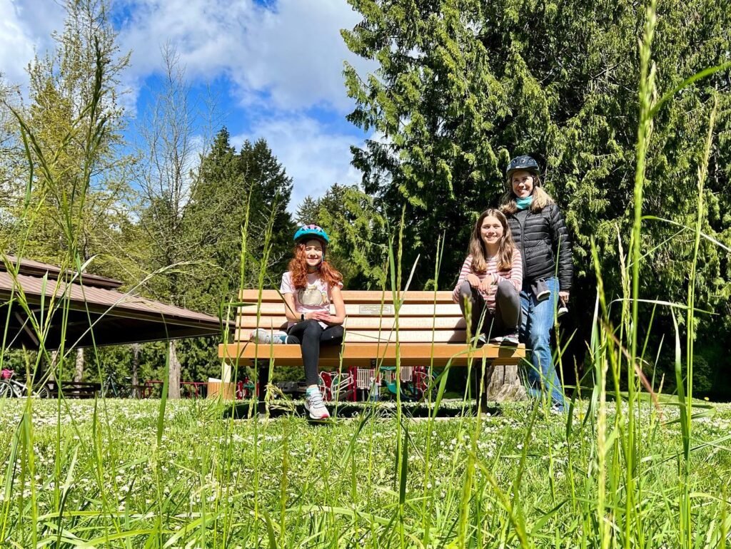 A family sits on a park bench in a grassy outdoor space under a blue sky. The setting includes tall trees and a rustic covered pavilion.