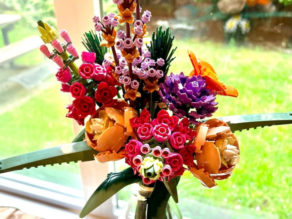A vibrant LEGO bouquet featuring red roses, pink lilies, and purple flowers displayed against a bright green outdoor background.