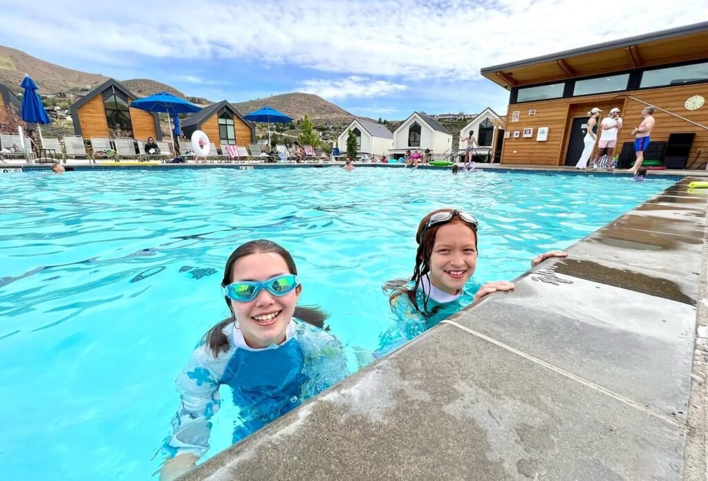 Two individuals wearing goggles swim in a turquoise geothermal pool, with thatched-roof cabins and hills in the background.