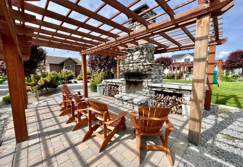 A wooden pergola structure provides shade over comfortable seating and a stone fire pit in a well-maintained garden.