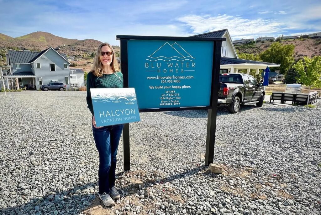 A traveler stands proudly in front of the Blu Water Cabins sign, holding a folder in a scenic gravel lot with a modern house in the background.