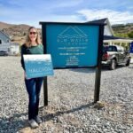 A traveler stands proudly in front of the Blu Water Cabins sign, holding a folder in a scenic gravel lot with a modern house in the background.