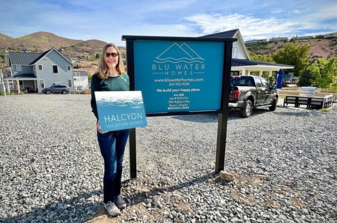 A traveler stands proudly in front of the Blu Water Cabins sign, holding a folder in a scenic gravel lot with a modern house in the background.