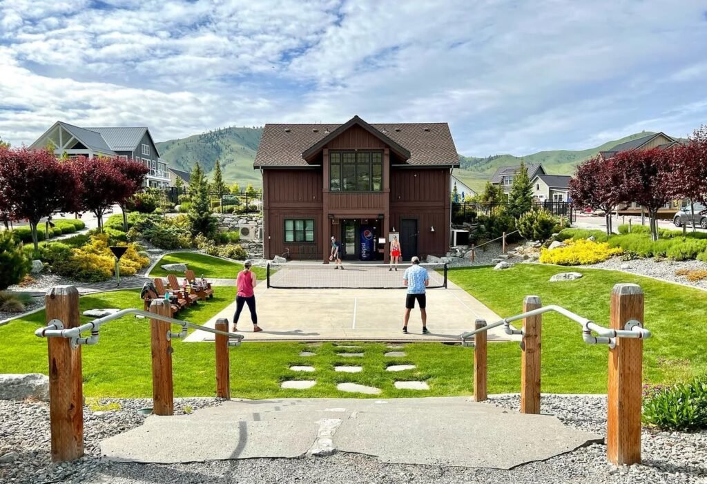 People playing pickleball on a concrete court with a large red wooden building and mountains in the background.