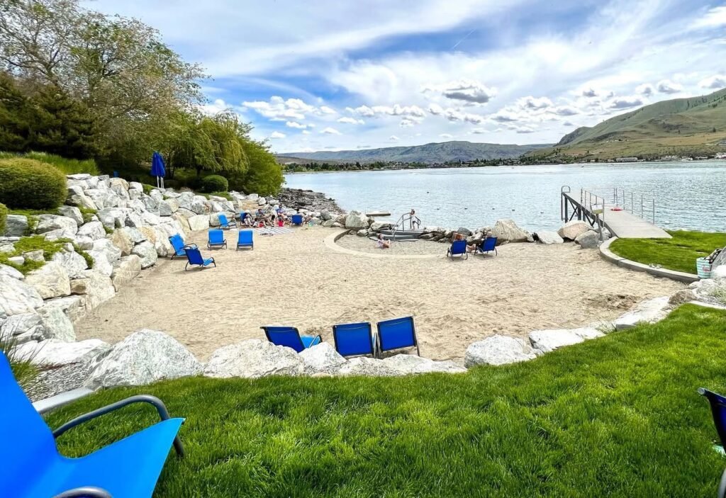 A scenic lakeside view featuring a private beach, blue lounge chairs, and a wooden dock under a partly cloudy sky.
