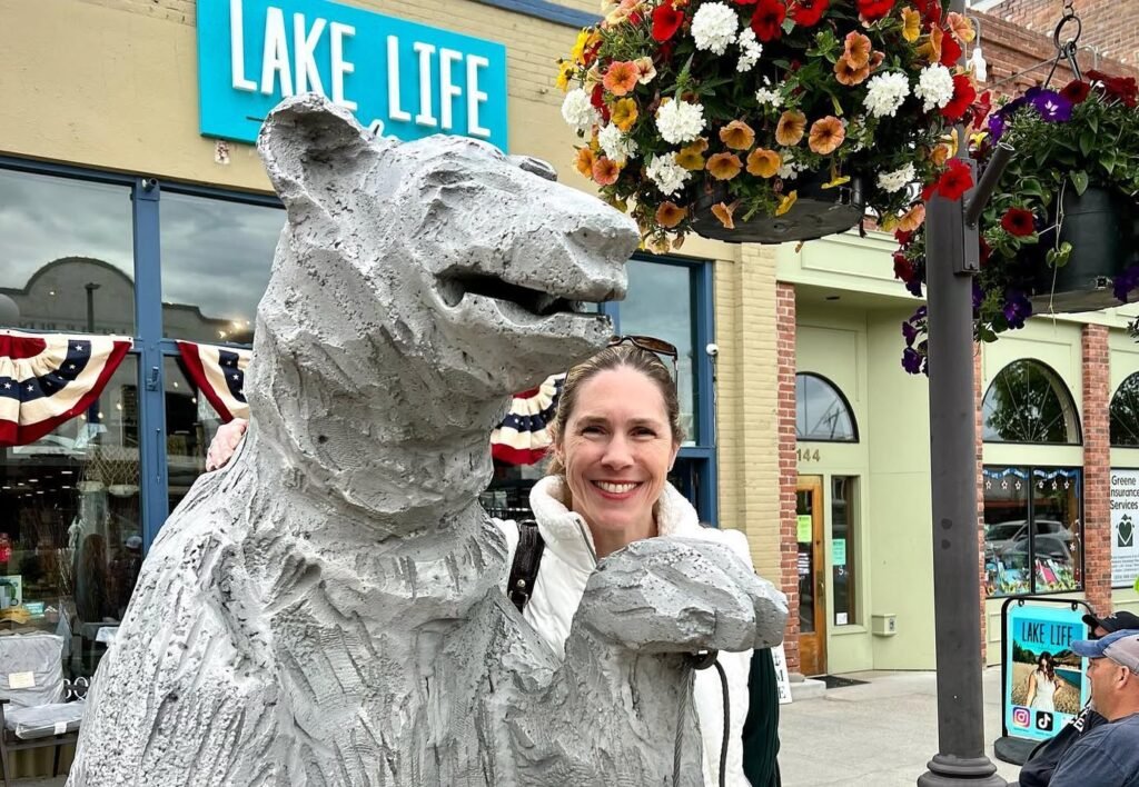 Smiling person posing next to a large mountain lion statue on the street, with hanging flower baskets and a sign in the background.
