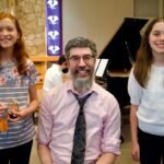 A family member and two young individuals capture happy moments at a formal gathering. Colorful banners and a grand piano create a festive backdrop.