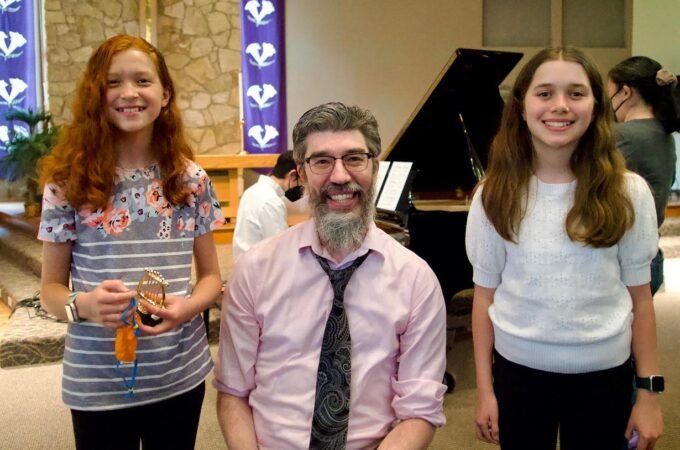 A family member and two young individuals capture happy moments at a formal gathering. Colorful banners and a grand piano create a festive backdrop.