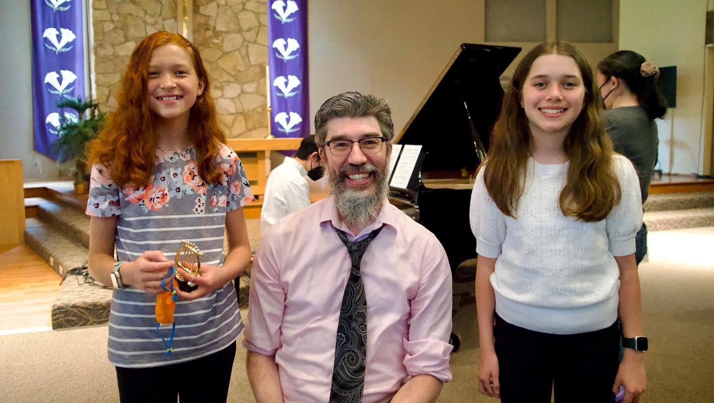Spring Piano Recital A family member and two young individuals capture happy moments at a formal gathering. Colorful banners and a grand piano create a festive backdrop.