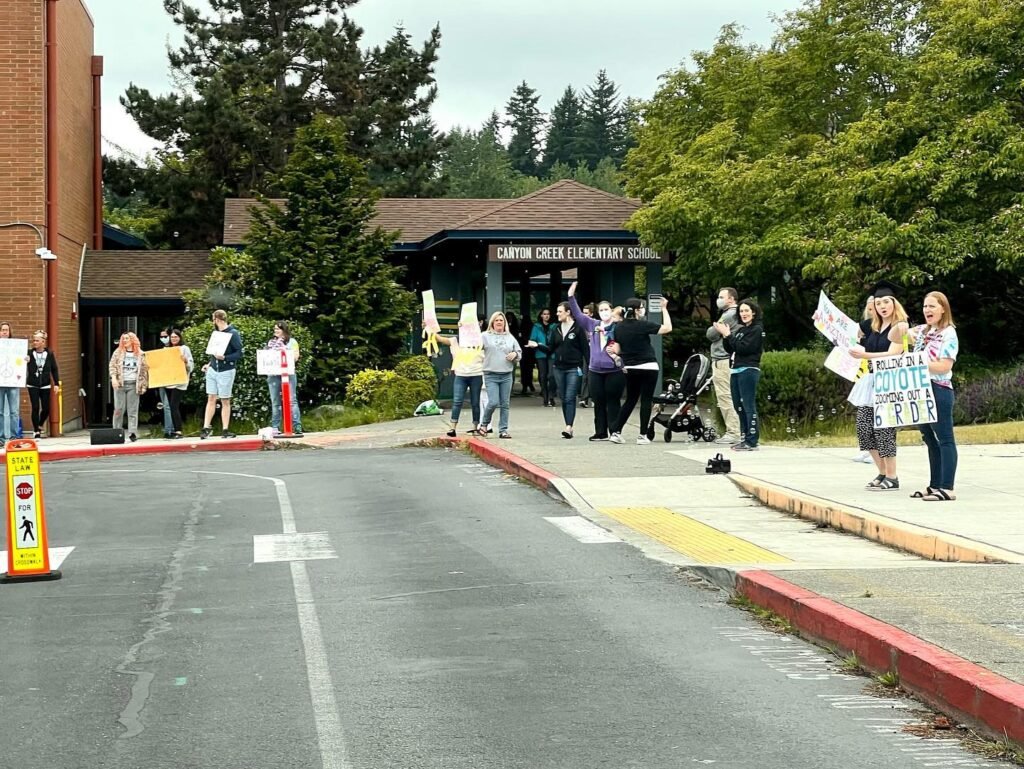 A group of people stands outside a brick building holding signs, with lush green trees in the background, reflecting a community gathering.