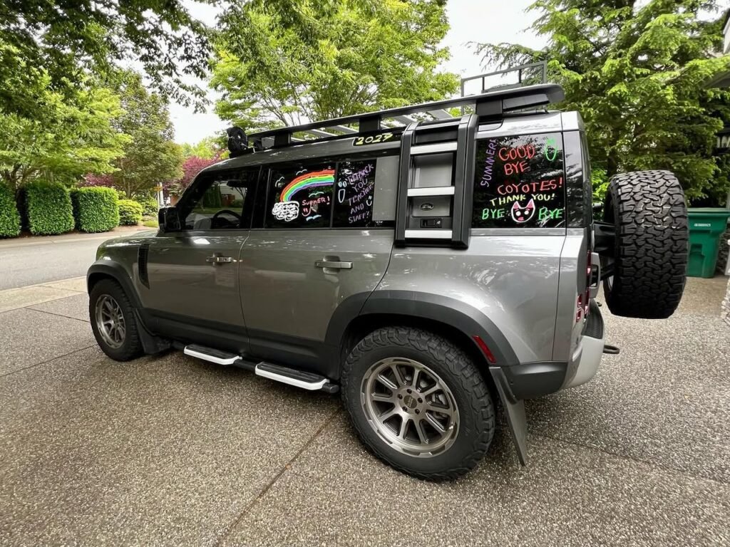 Custom gray SUV with a ladder and roof rack parked outside a house with rainbow decals.