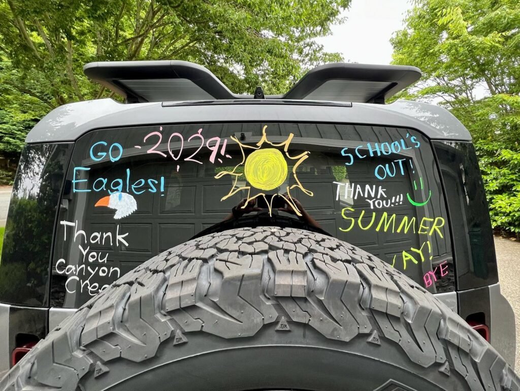 A car's rear window displays "Go Eagles! 2021", "School's Out", and "Thank You Canyon Creek", paired with a large off-road tire and summer greetings amid green foliage.
