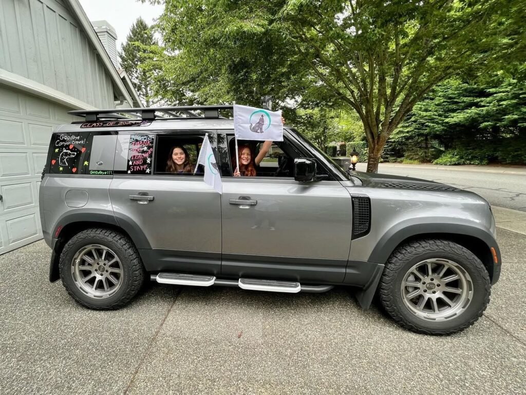 A family waves from a customized gray SUV at a garage. The Pacific Northwest is visible in the lush background.