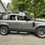 A family waves from a customized gray SUV at a garage. The Pacific Northwest is visible in the lush background.