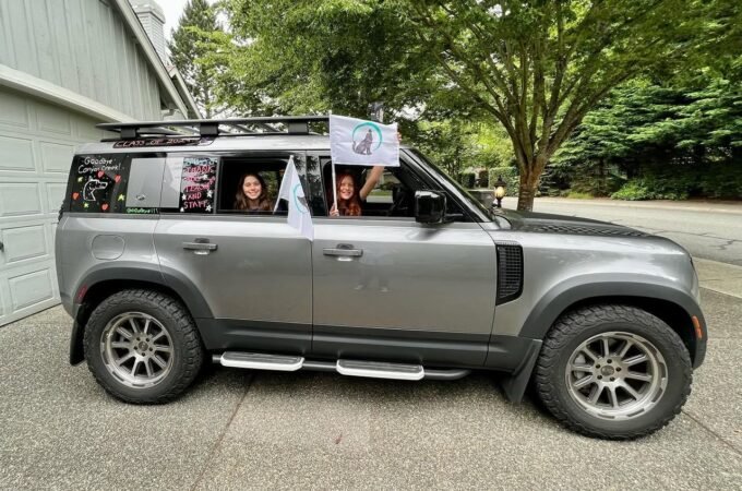 A family waves from a customized gray SUV at a garage. The Pacific Northwest is visible in the lush background.
