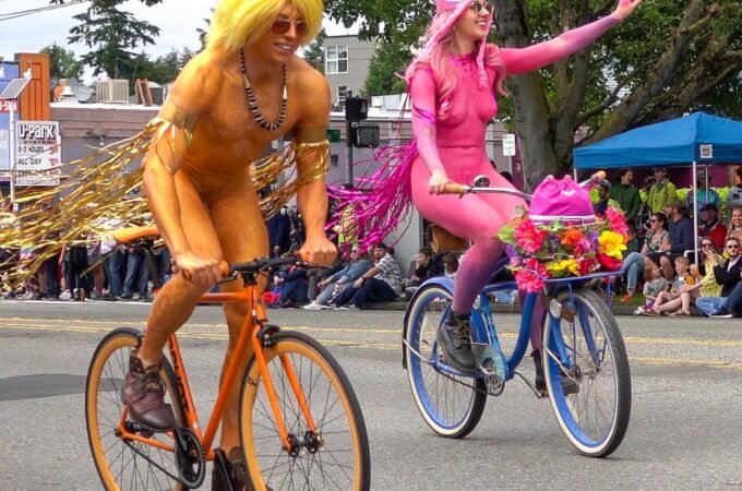 Two people in vibrant costumes riding bicycles on a public street. Spectators line the road, watching the parade in the Pacific Northwest.