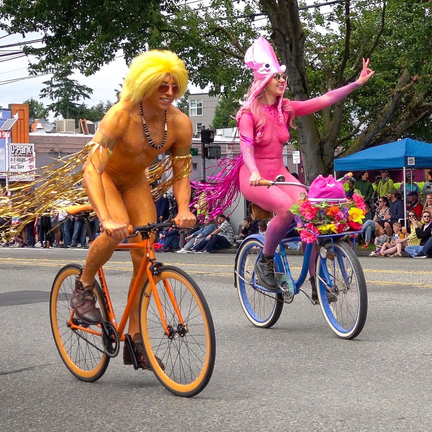 Two people in vibrant costumes riding bicycles on a public street. Spectators line the road, watching the parade in the Pacific Northwest.