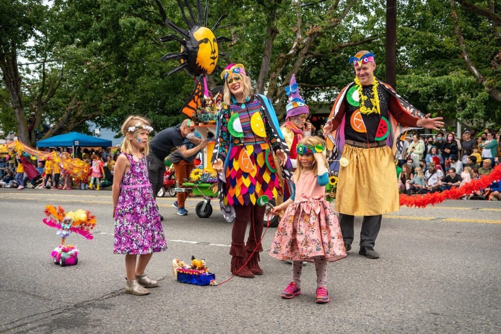 Participants in vibrant costumes parade through a lush park, with children posing for photos in front of a large whimsical puppet decoration.