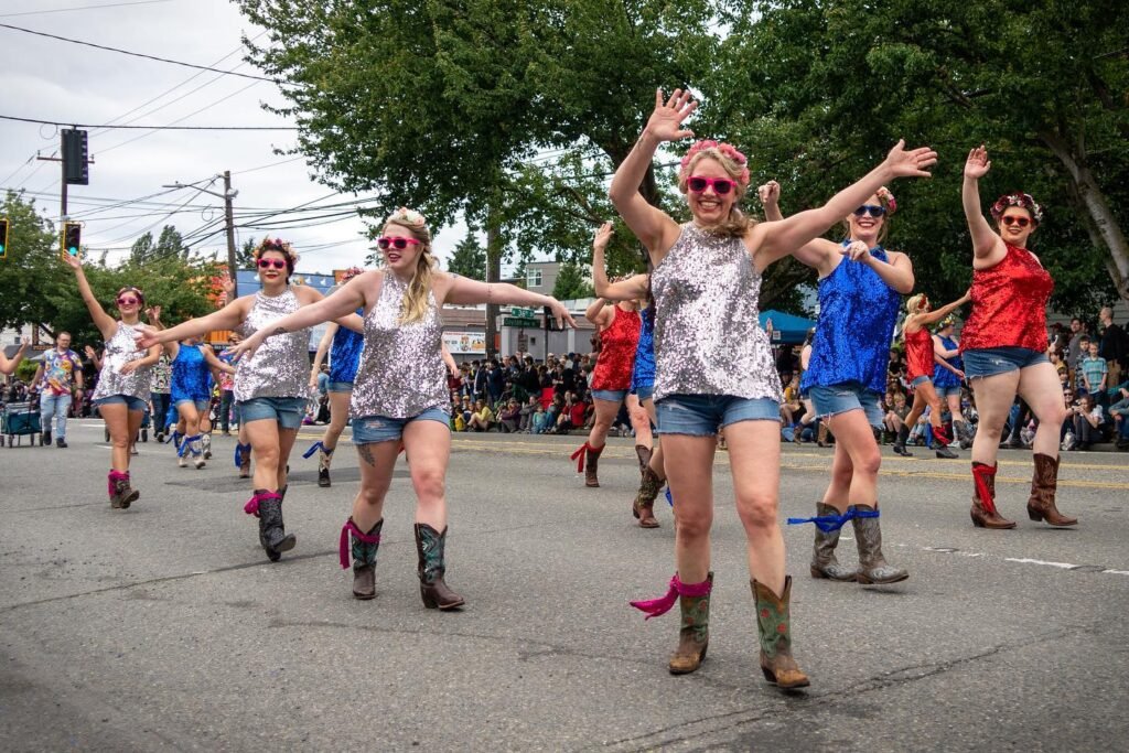 A group of dancers in colorful outfits and cowboy boots perform on a city street during a daytime festival.