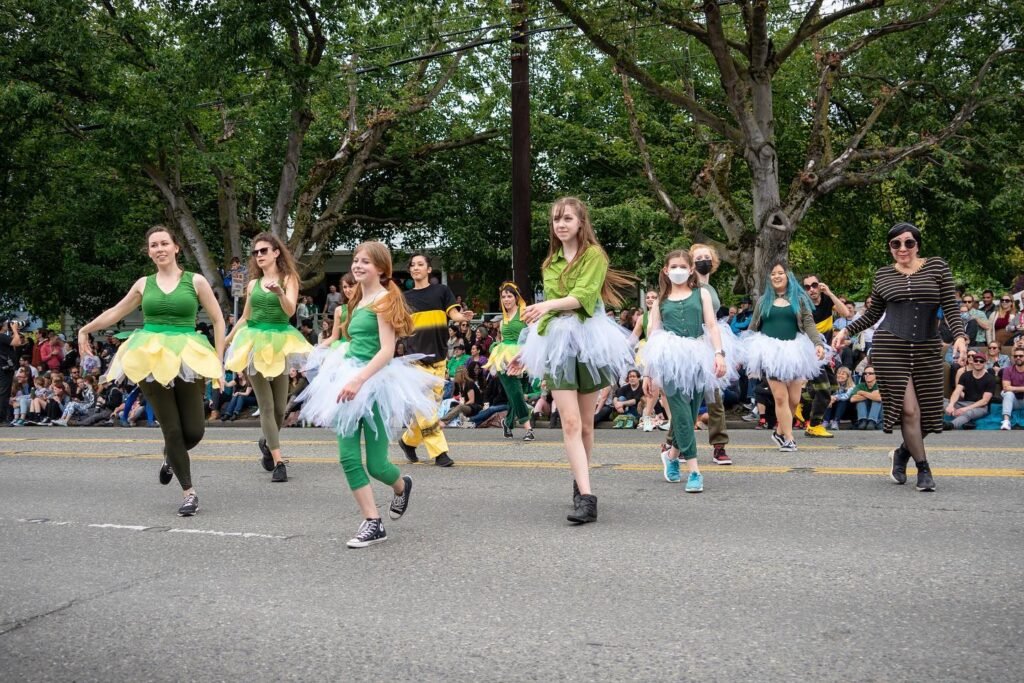 Performers in vibrant green and yellow dresses march down a paved street lined with tall trees. Spectators line the sidewalks watching the festive parade.
