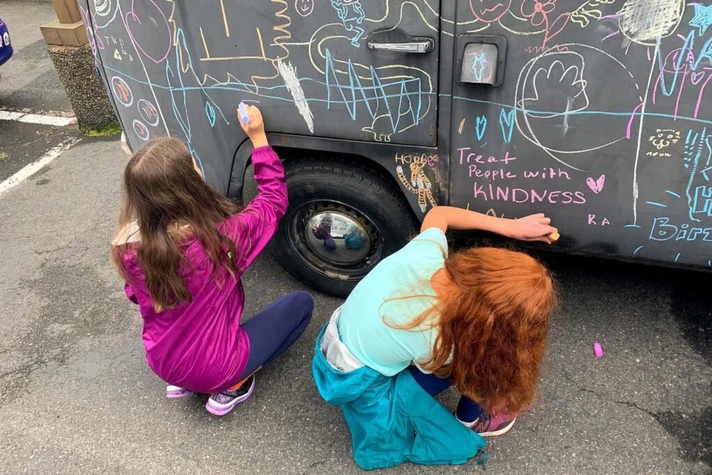 Two kids drawing colorful chalk art on a dark van that reads 'treat people with KINDNESS'.