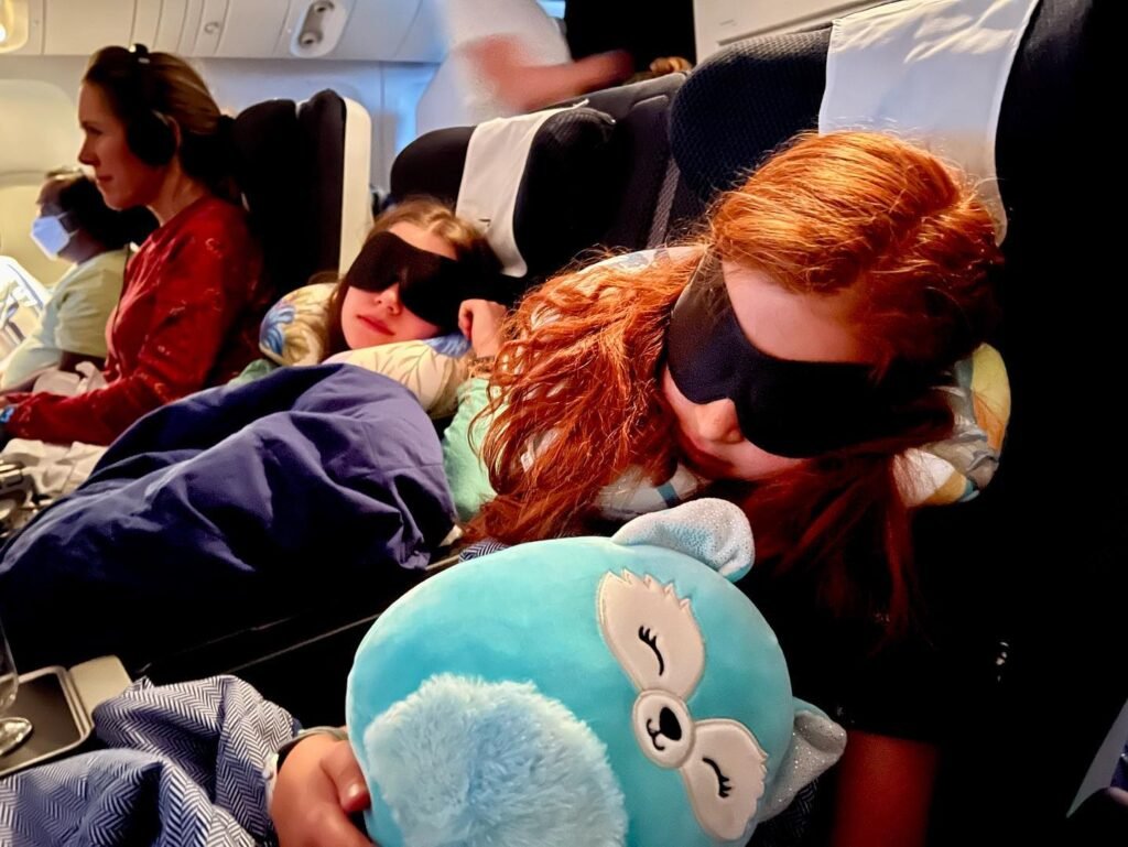 Passengers resting in airplane seats with eye masks, one holding a blue plush toy near the window.