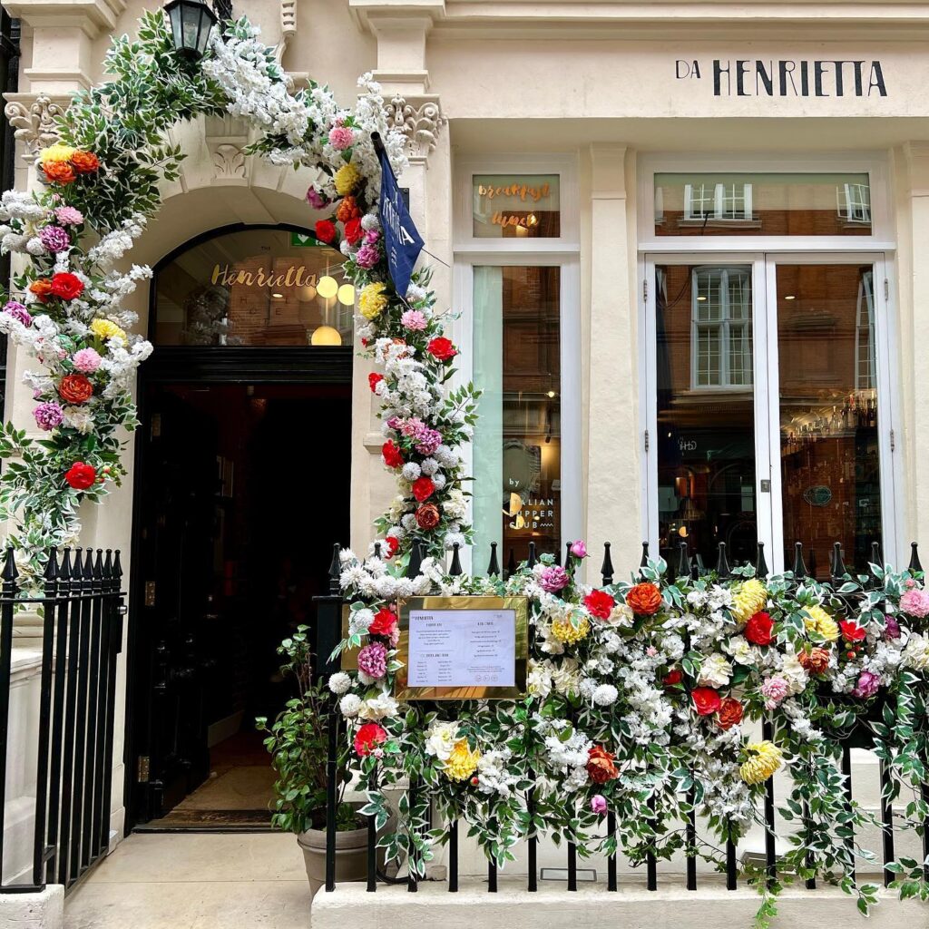 A decorative entrance to La Henrietta featuring a lush floral arch over the door and window displays.