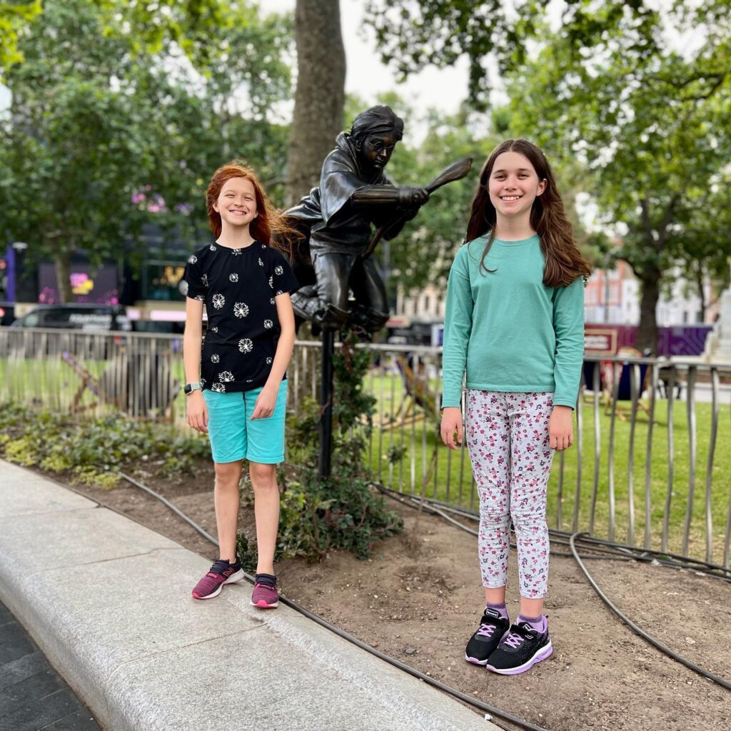 Two young girls posing beside a bronze statue in a tree-lined urban park.