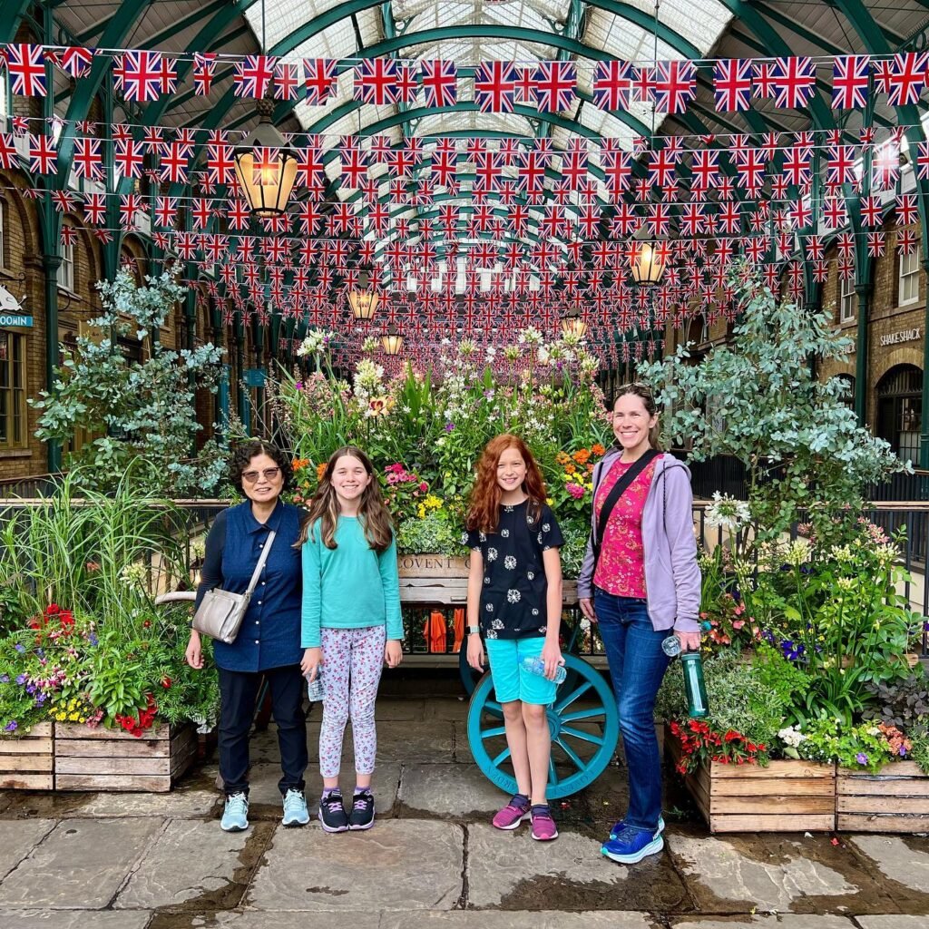 A family poses under a canopy of British flags in a vibrant market garden setting. Lush foliage and colorful flowers complete the festive scene.