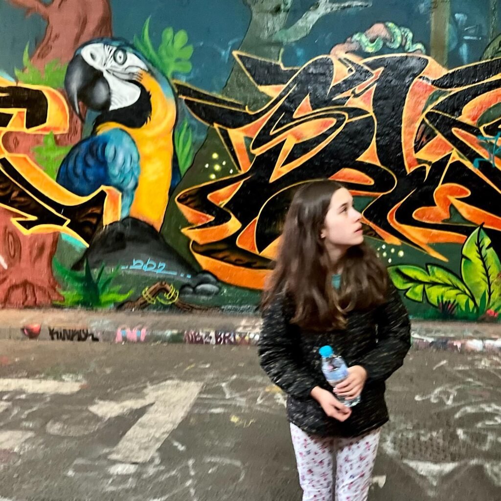 A young woman stands in front of a vibrant street art mural featuring a blue and yellow macaw parrot.