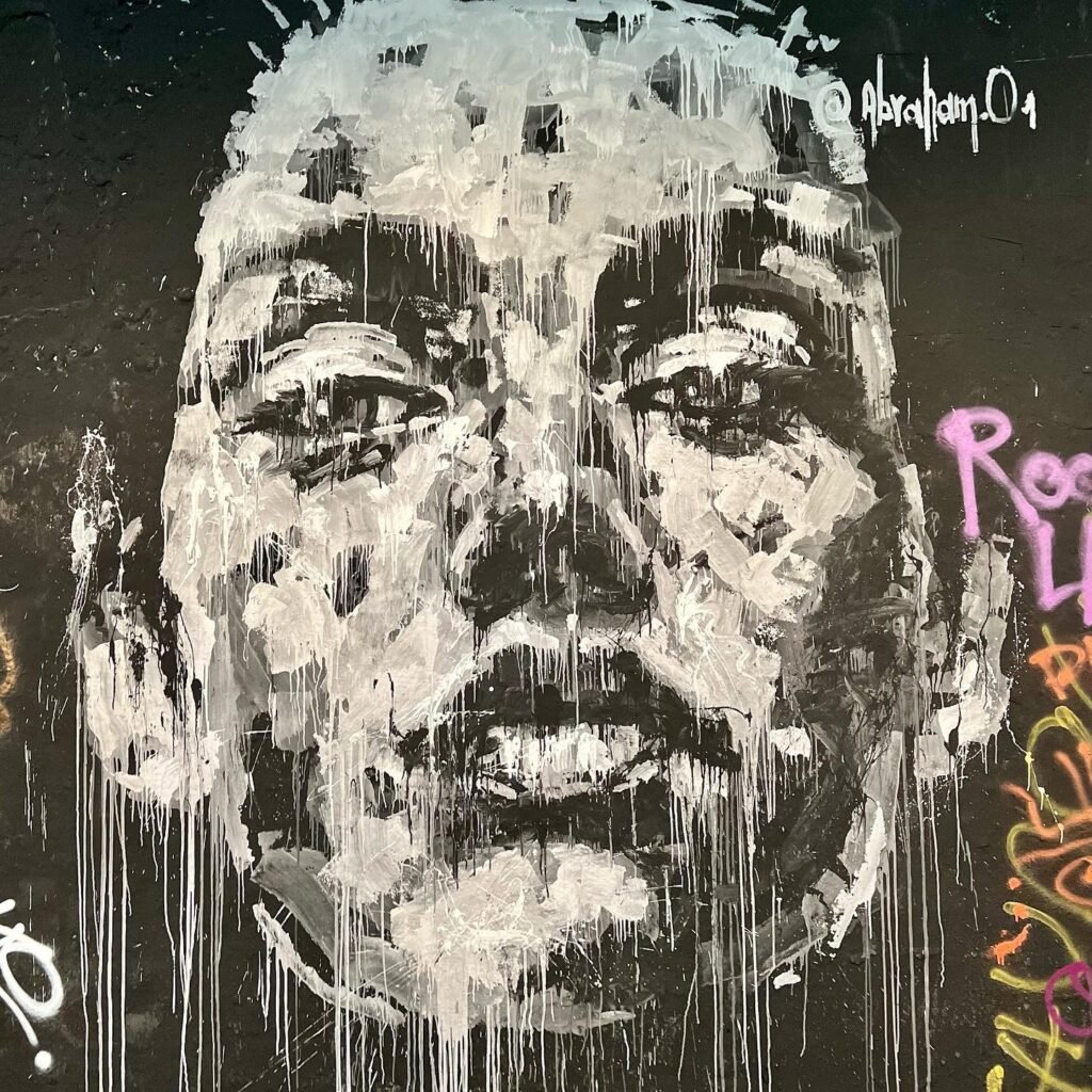 A monochromatic stencil portrait with dripping paint effects on a dark background, accompanied by colorful secondary graffiti tags.