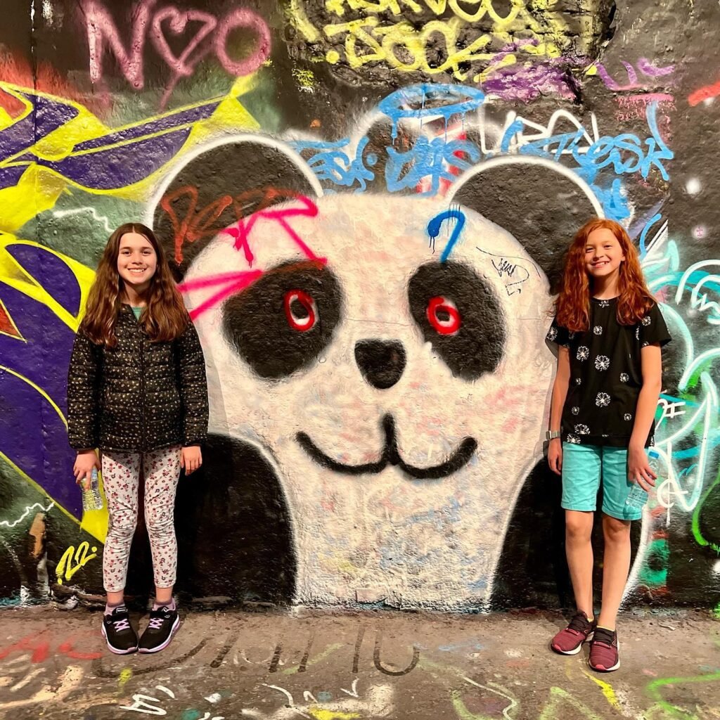 Two girls standing side by side in front of a large, vibrant panda mural with spray paint graffiti on the wall.