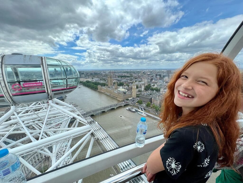 A person with red hair smiles inside a London Eye pod with a wide view of the city and river below.