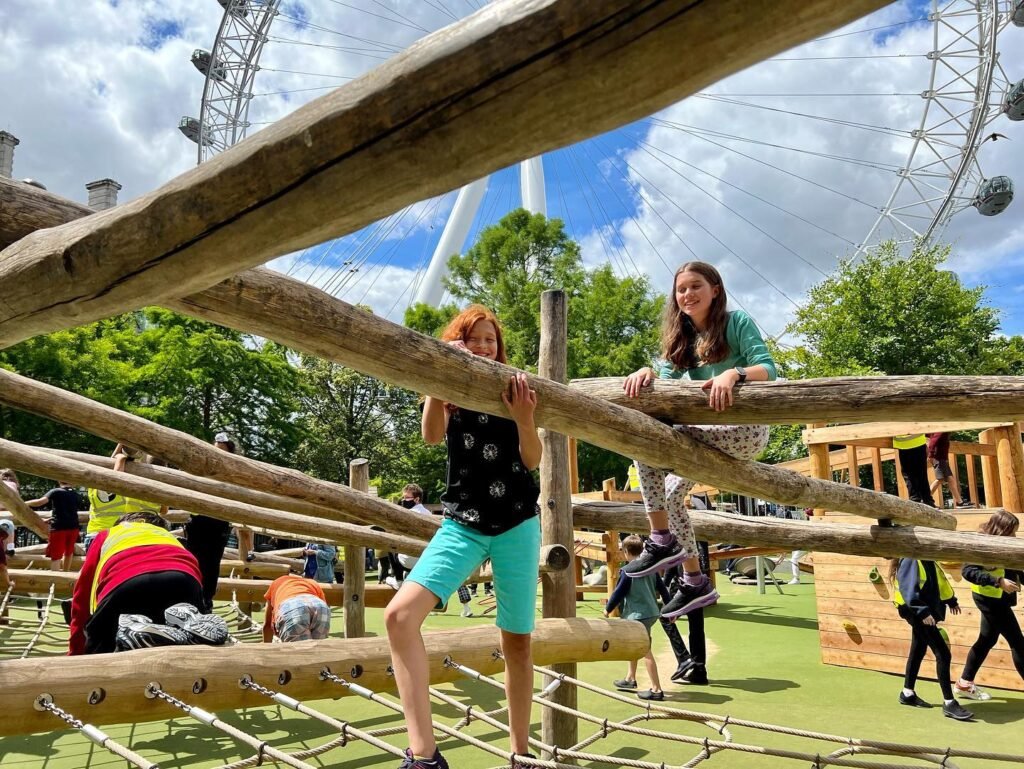 A wooden play structure with children climbing and exploring. A large Ferris wheel is visible in the background.