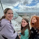 Three people smiling and posing for a photo on a high observation deck with a view of London. Part of the London Eye is visible in the background.