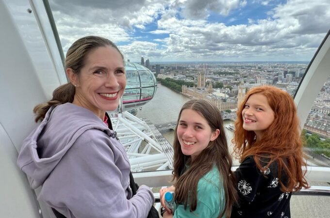 Three people smiling and posing for a photo on a high observation deck with a view of London. Part of the London Eye is visible in the background.