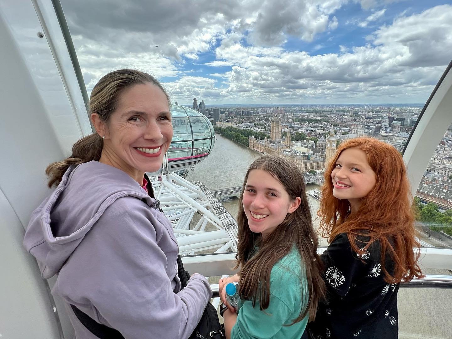 London Eye Three people smiling and posing for a photo on a high observation deck with a view of London. Part of the London Eye is visible in the background.