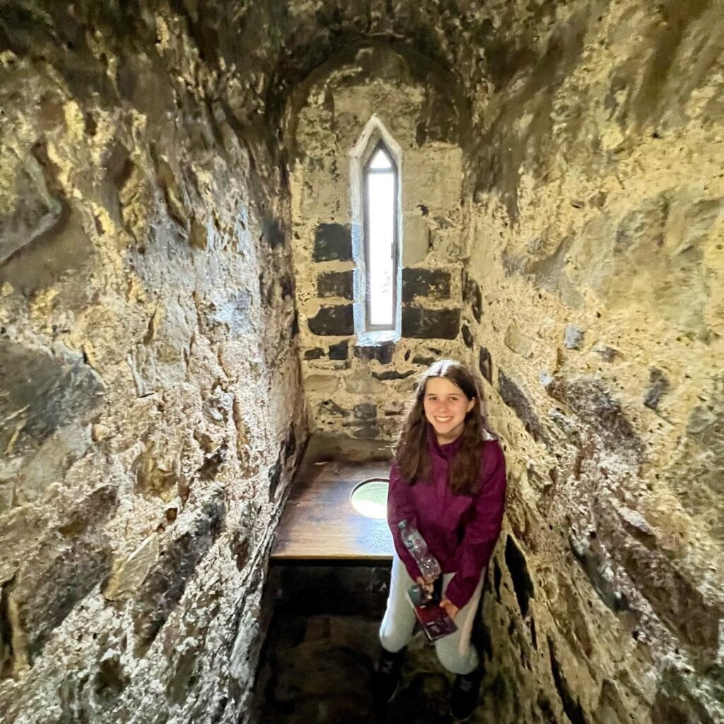 A young girl stands inside a narrow, historic stone corridor with a gothic arch. Natural light streams in through a small, narrow window in the background.