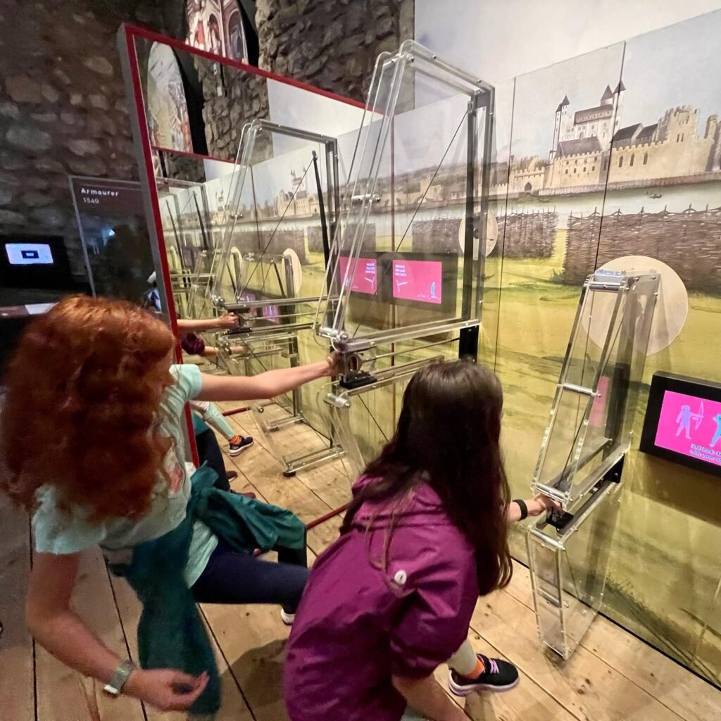 Tourists interacting with a mirrored, lens-based exhibit set against a mural of the Tower of London, London, UK.
